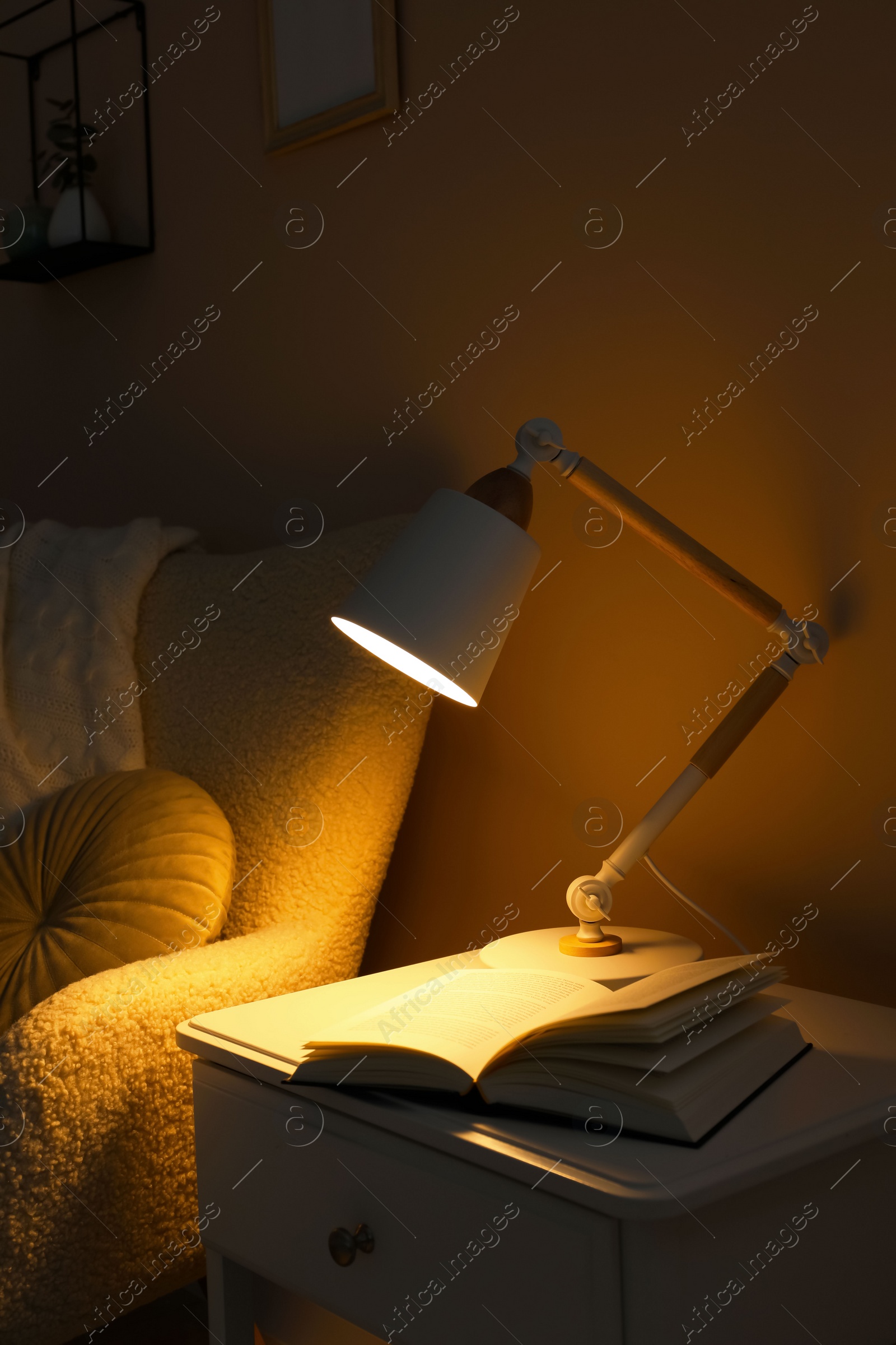 Photo of Stylish modern desk lamp and open book on white cabinet near armchair in dark room