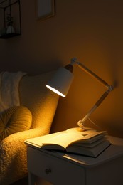 Photo of Stylish modern desk lamp and open book on white cabinet near armchair in dark room