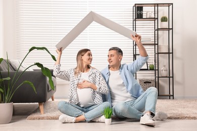 Young family housing concept. Pregnant woman with her husband sitting under cardboard roof on floor at home
