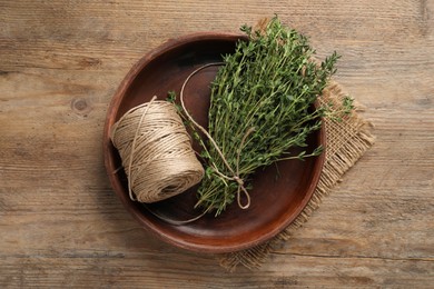 Photo of Bunch of aromatic thyme and twine in bowl on wooden table, top view
