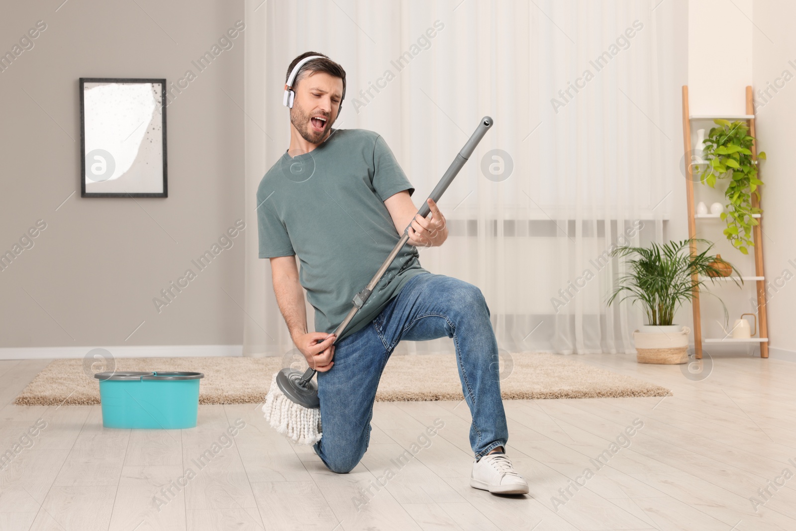 Photo of Enjoying cleaning. Happy man in headphones with mop singing while tidying up at home