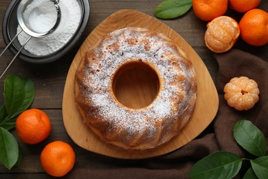 Photo of Homemade yogurt cake with tangerines, powdered sugar and green leaves on wooden table, flat lay