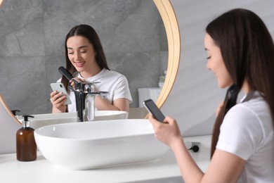 Photo of Beautiful young woman with hair iron using smartphone in bathroom. Internet addiction