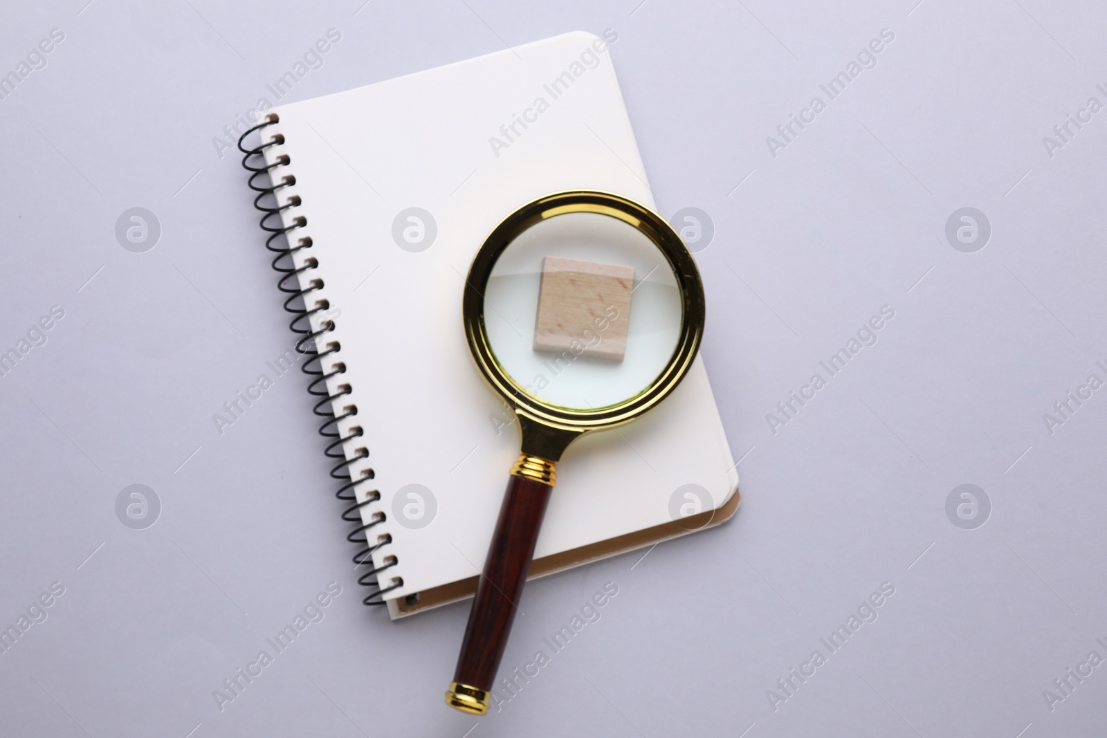Photo of Magnifying glass over piece of wood and notebook on light grey background, top view