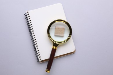 Magnifying glass over piece of wood and notebook on light grey background, top view
