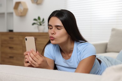 Photo of Young woman having video chat via smartphone and blowing kiss on sofa in living room