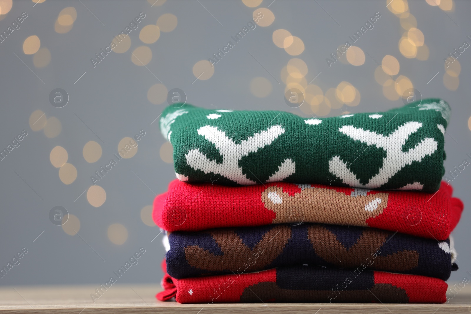 Photo of Stack of different Christmas sweaters on table against grey background with blurred lights. Space for text