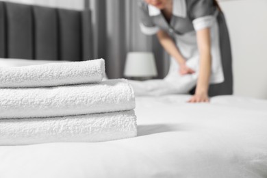 Photo of Young maid making bed in hotel room, focus on stack of clean towels