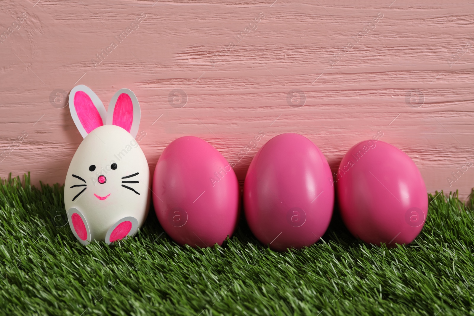 Photo of Bright eggs and white one as Easter bunny on green grass against pink wooden background