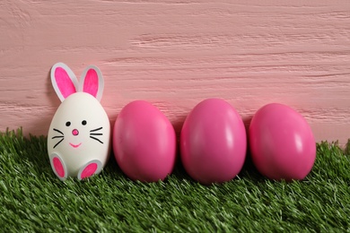 Photo of Bright eggs and white one as Easter bunny on green grass against pink wooden background