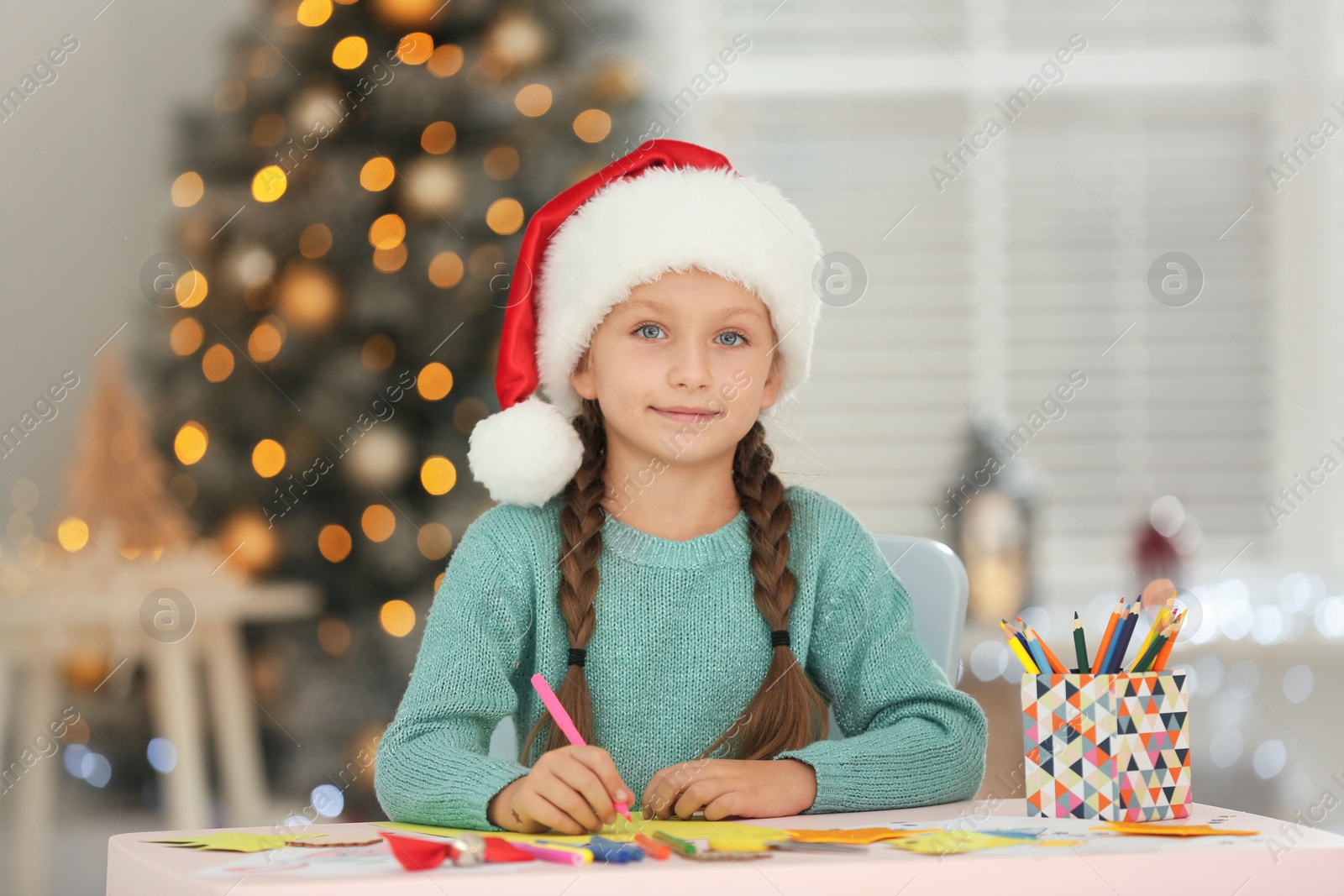 Photo of Little child in Santa hat drawing at table indoors. Christmas season