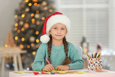 Photo of Little child in Santa hat drawing at table indoors. Christmas season