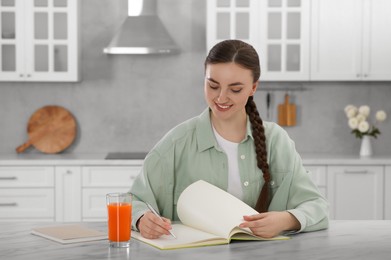 Photo of Beautiful young woman writing in notebook at white marble table indoors