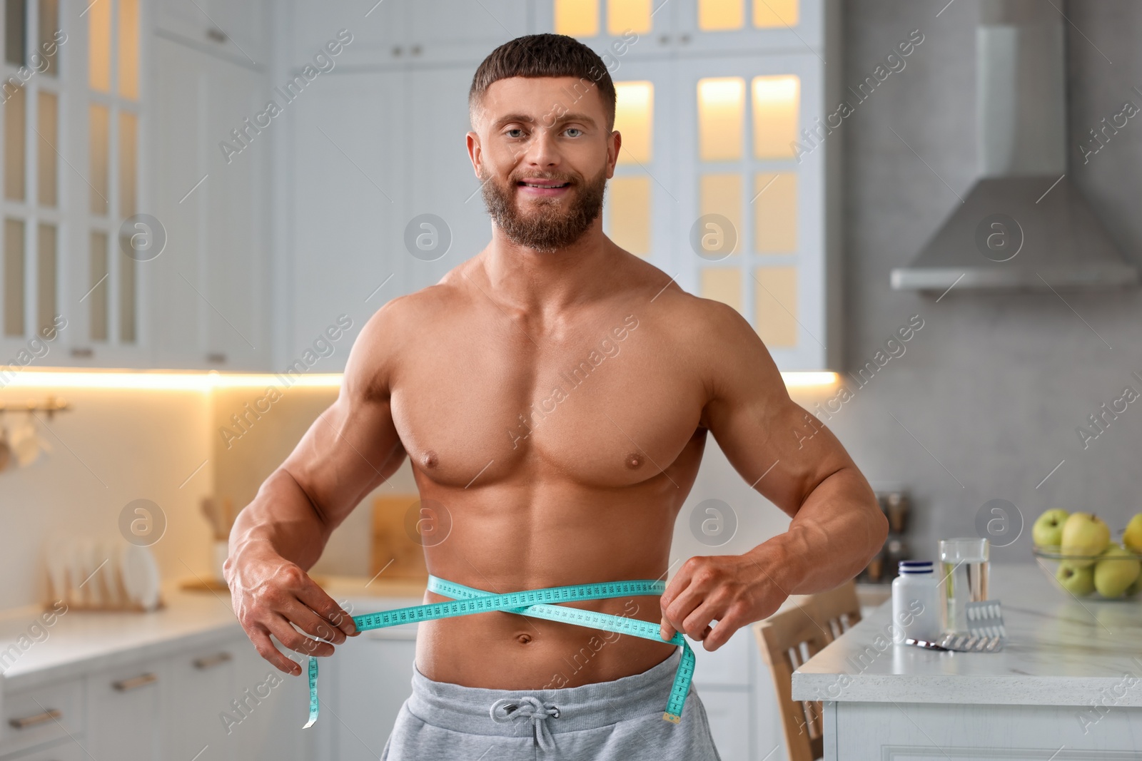 Photo of Athletic young man measuring his waist with tape in kitchen. Weight loss