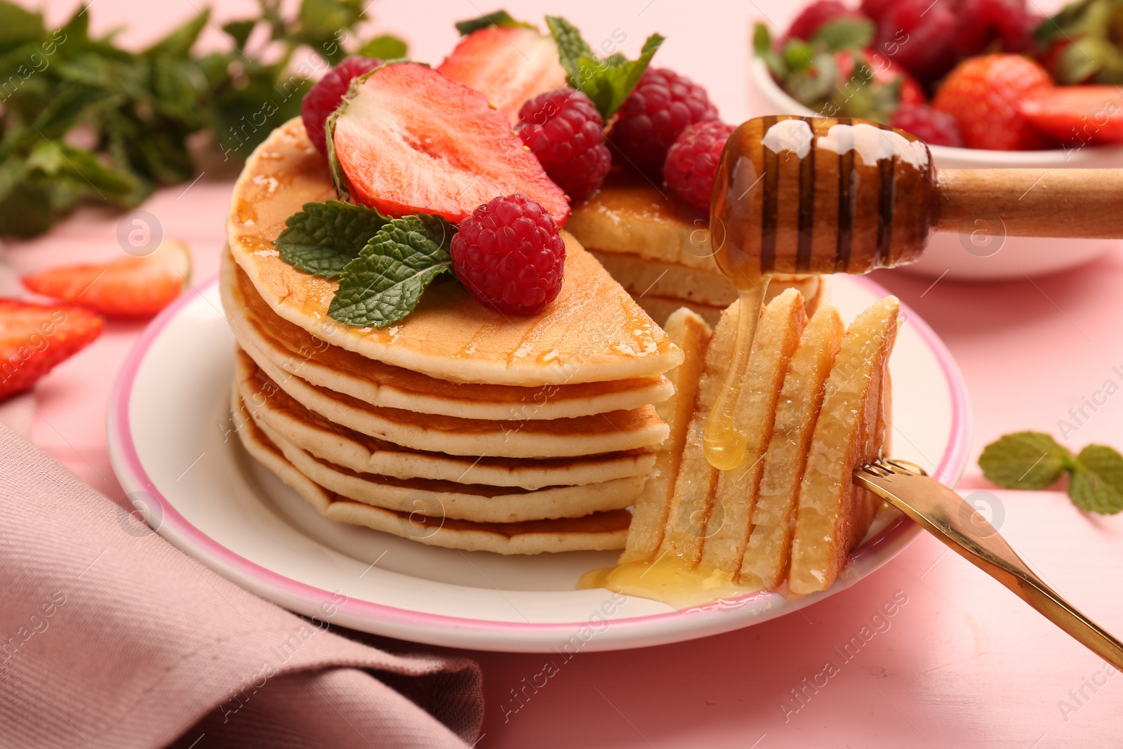 Photo of Dripping honey onto delicious pancakes with fresh berries on pink table, closeup