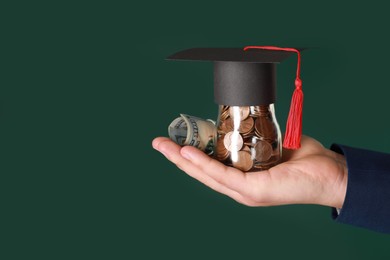 Photo of Man holding glass jar of coins, dollar banknotes and graduation cap against green background, closeup with space for text. Scholarship concept