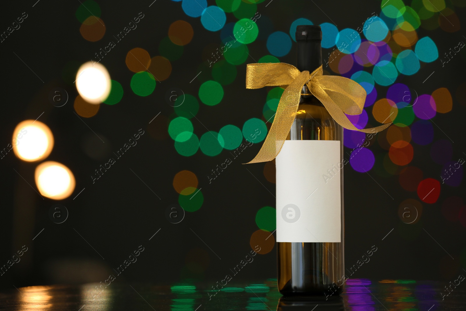 Photo of Bottle of wine with bow-knot on table against blurred lights, space for text. Bokeh effect