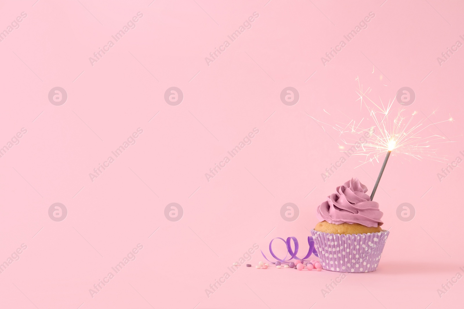 Photo of Birthday cupcake with burning sparkler, streamer and sprinkles on pink background. Space for text