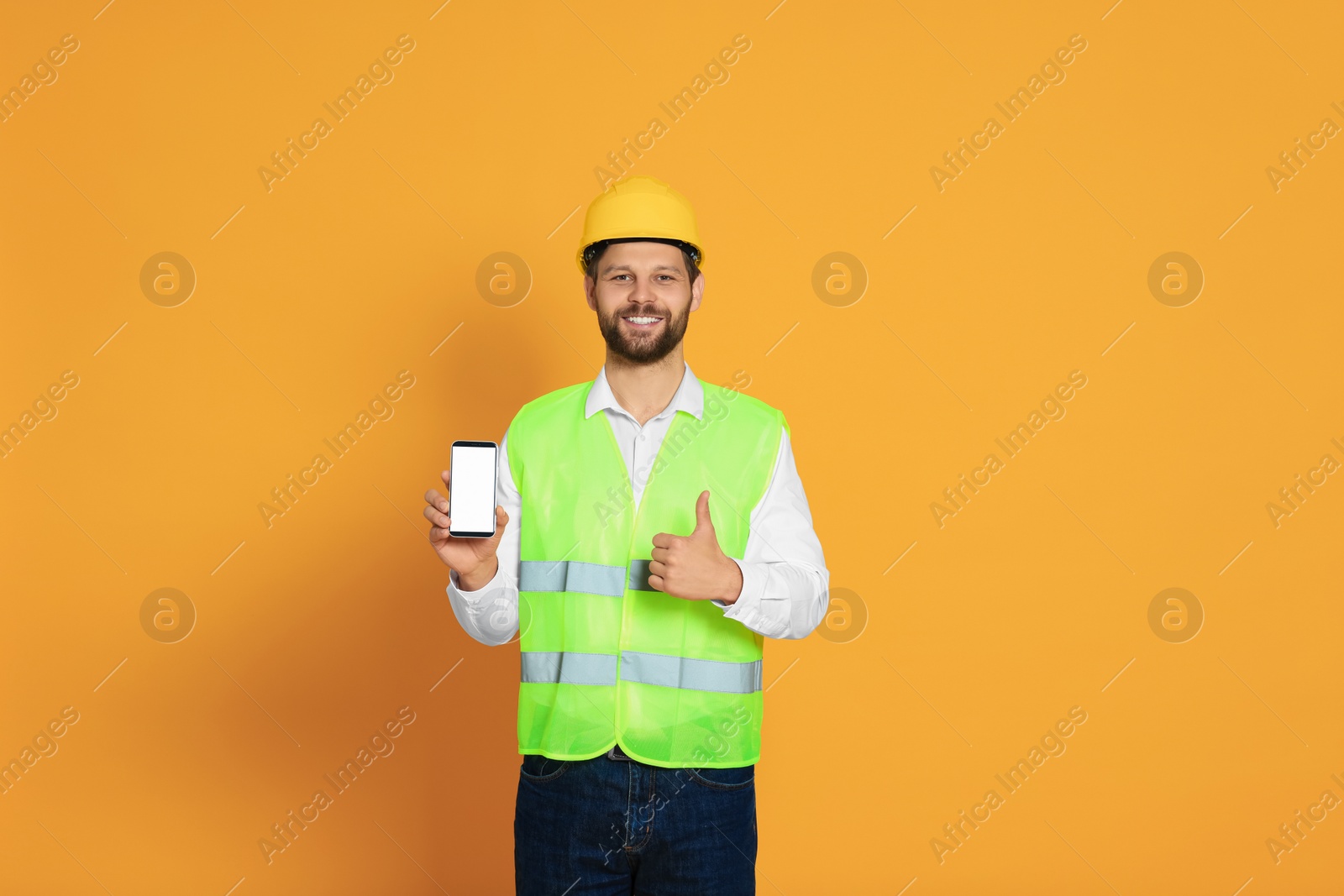 Photo of Man in reflective uniform showing smartphone and thumbs up on orange background