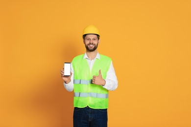 Photo of Man in reflective uniform showing smartphone and thumbs up on orange background