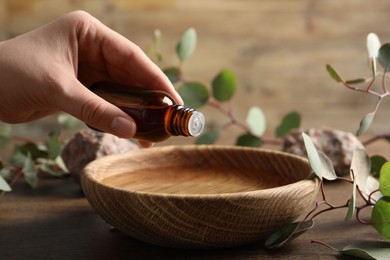 Photo of Woman dripping eucalyptus essential oil from bottle into bowl at wooden table, closeup