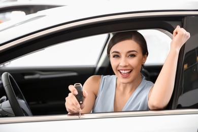 Young woman with key sitting in driver's seat of new car at salon