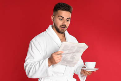 Photo of Young man in bathrobe with cup of coffee reading newspaper on red background