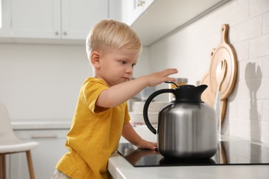Photo of Curious little boy playing with kettle on electric stove in kitchen