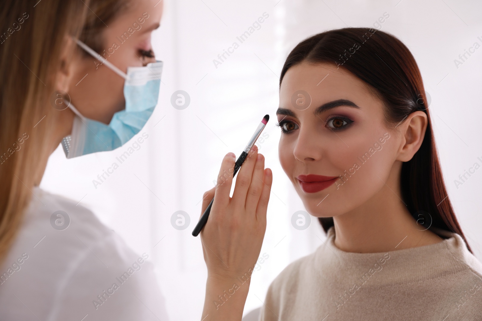 Photo of Makeup artist in protective mask working with woman indoors. Preventive measures during COVID-19 pandemic