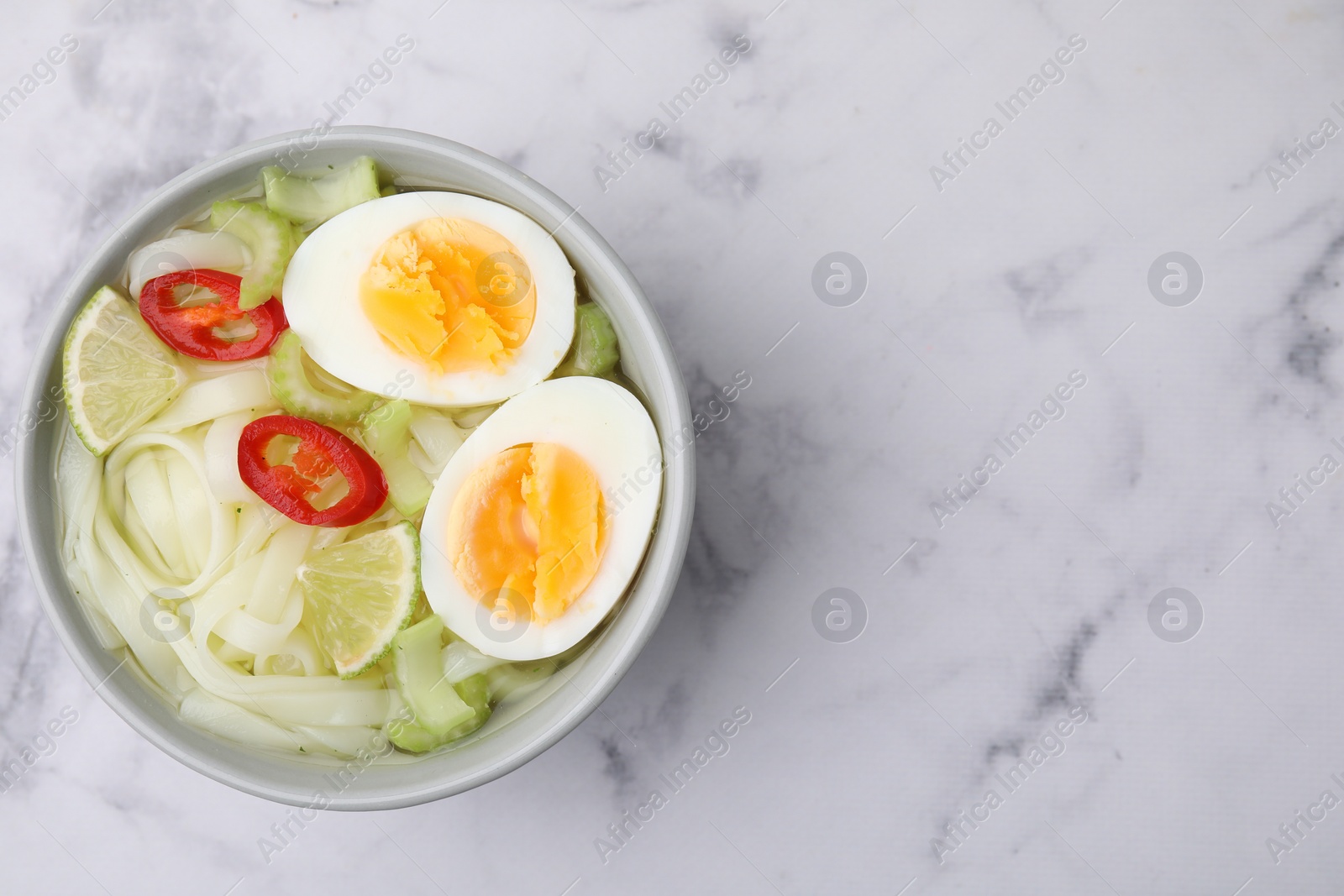 Photo of Bowl of delicious rice noodle soup with celery and egg on white marble table, top view. Space for text