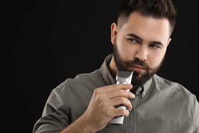 Handsome young man trimming beard on black background