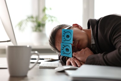 Man with fake eyes painted on sticky notes snoozing at workplace in office