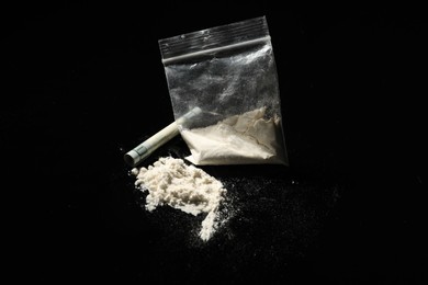 Photo of Drug addiction. Plastic bag with cocaine and rolled dollar banknote on black background