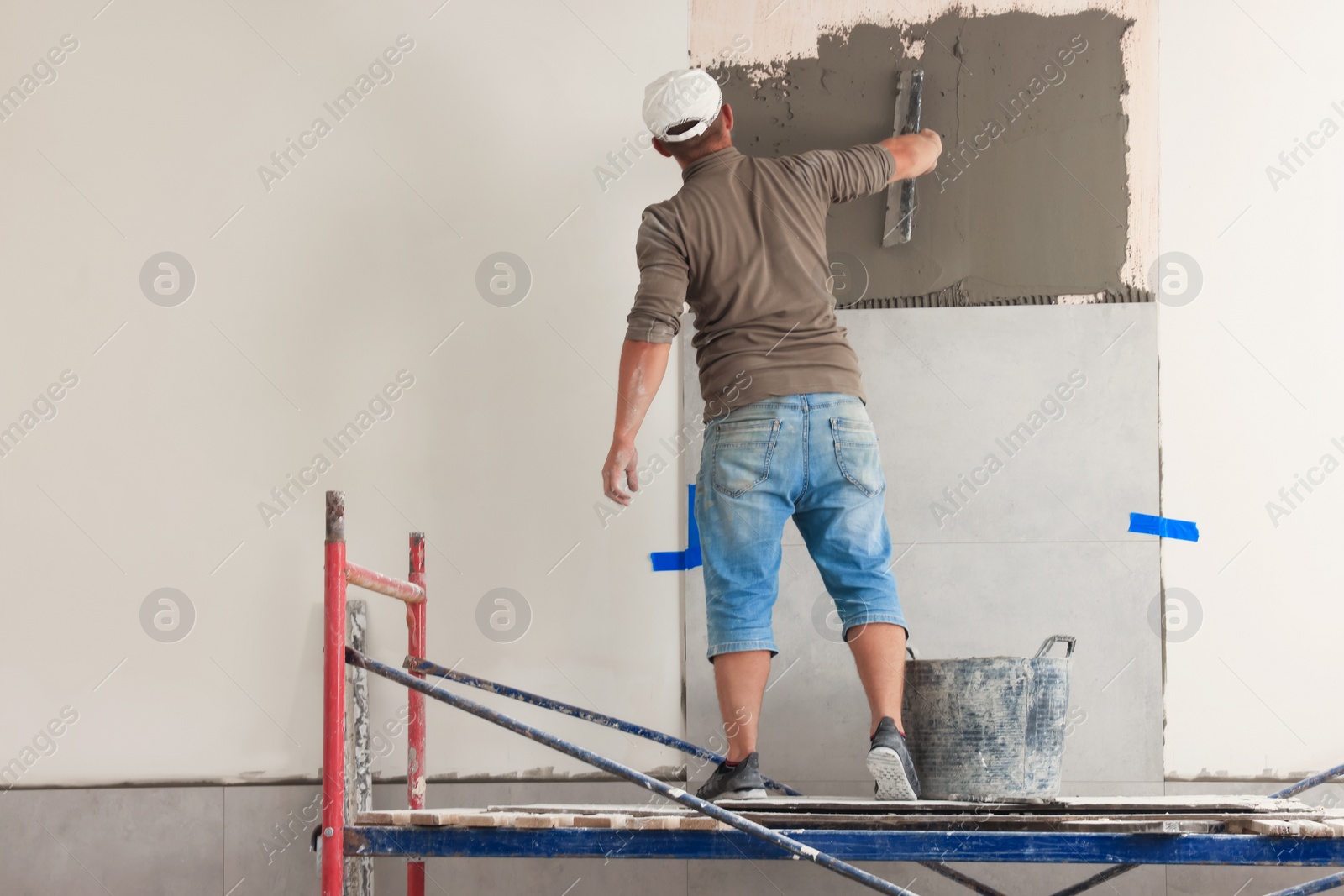 Photo of Worker spreading adhesive mix over wall for tile installation indoors, back view