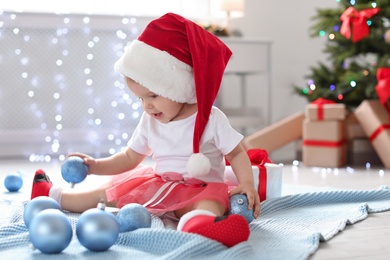 Photo of Cute baby in festive costume playing with Christmas decor on floor at home