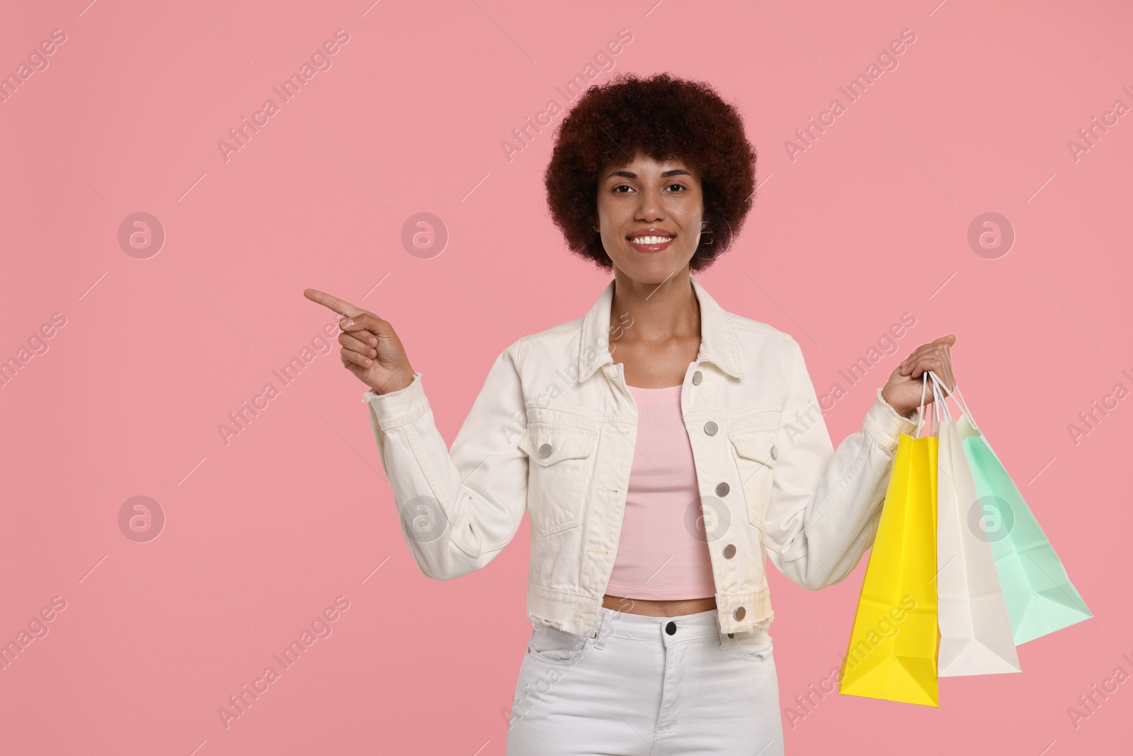 Photo of Happy young woman with shopping bags pointing at something on pink background. Space for text