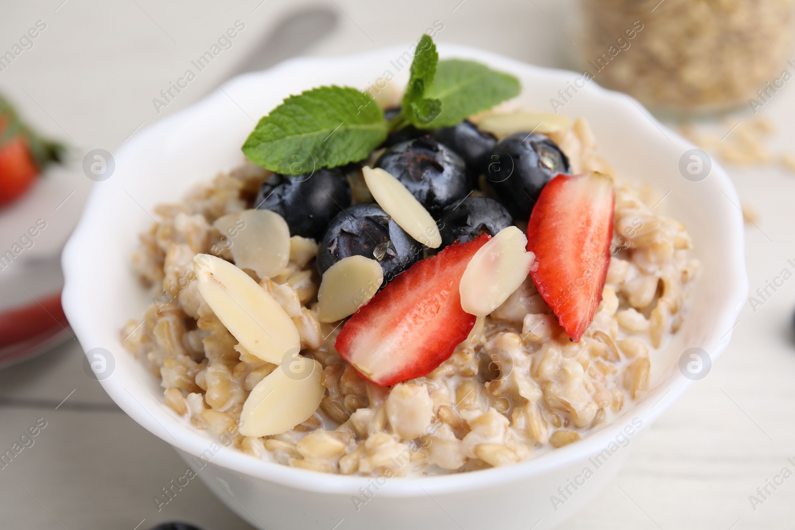 Photo of Tasty oatmeal with strawberries, blueberries and almond petals in bowl on white wooden table, closeup