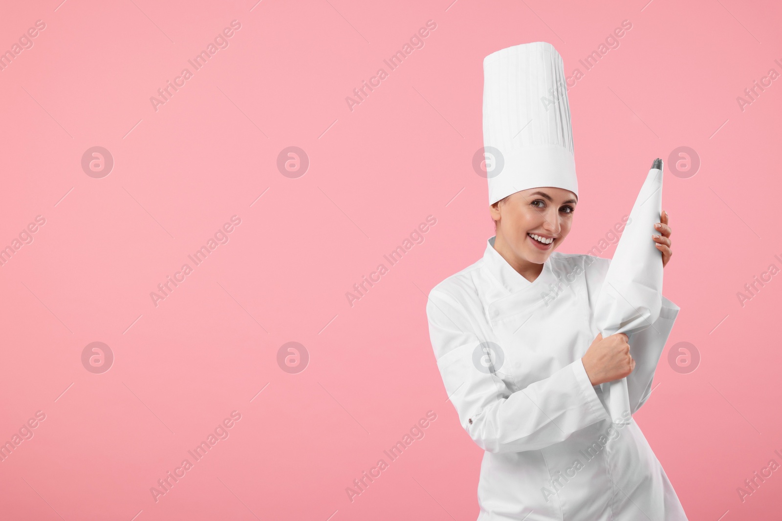 Photo of Happy professional confectioner in uniform holding piping bag on pink background. Space for text