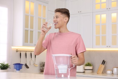 Happy man drinking clear water near filter jug at table in kitchen