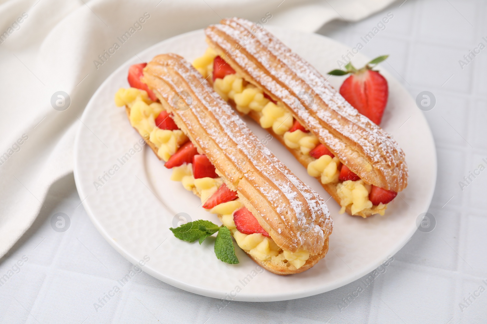 Photo of Delicious eclairs filled with cream, strawberries and mint on white tiled table