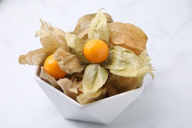 Photo of Ripe physalis fruits with calyxes in bowl on white marble table