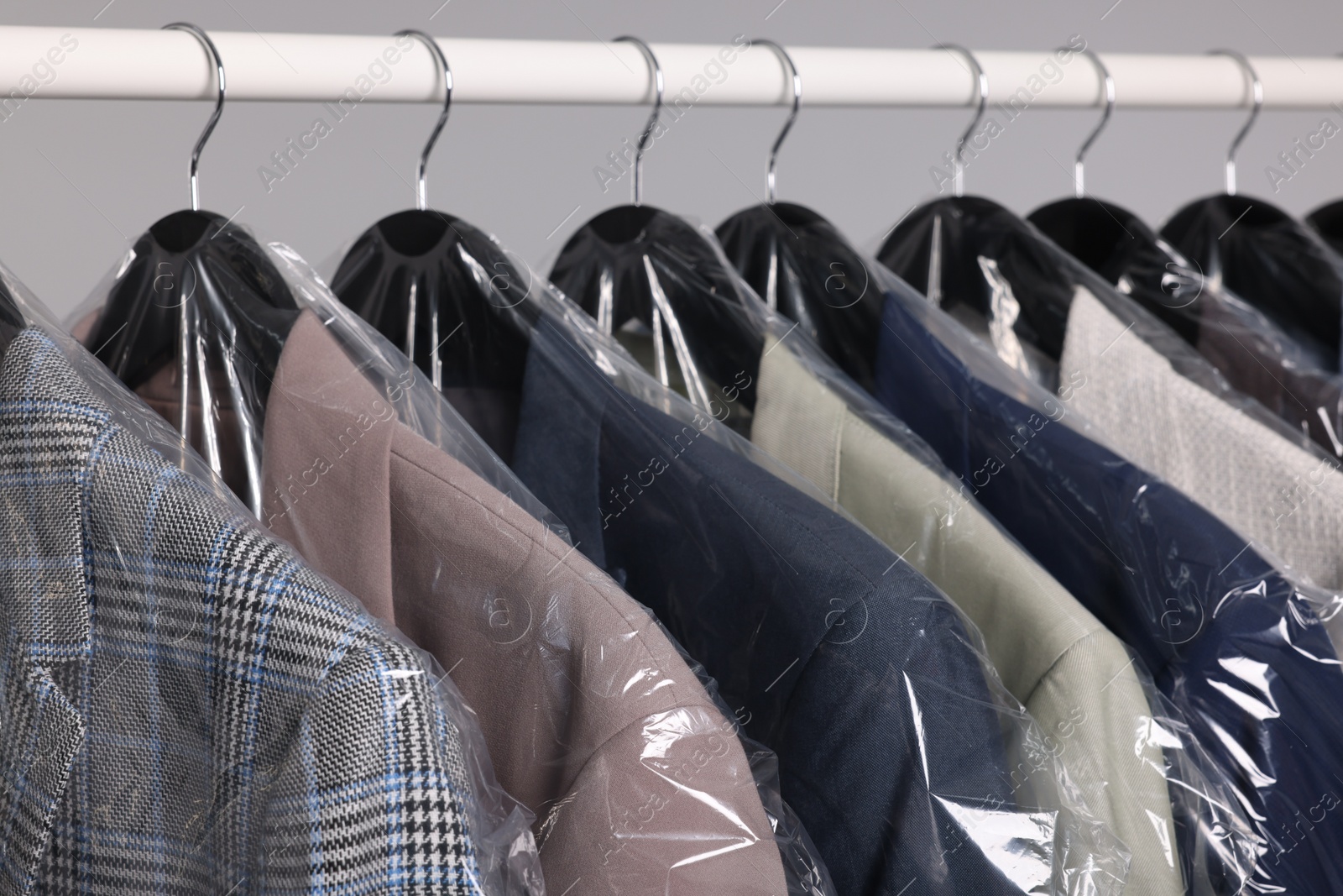 Photo of Dry-cleaning service. Many different clothes in plastic bags hanging on rack against grey background, closeup