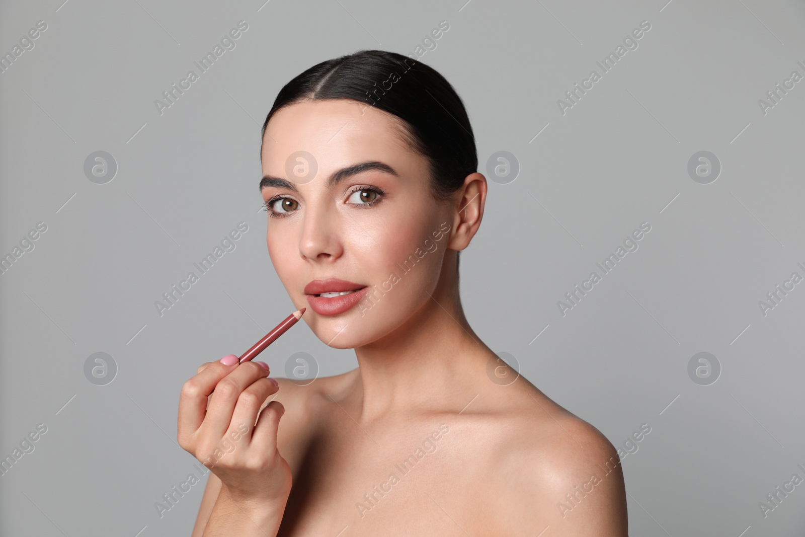 Photo of Pretty young woman applying beautiful nude lip pencil on grey background