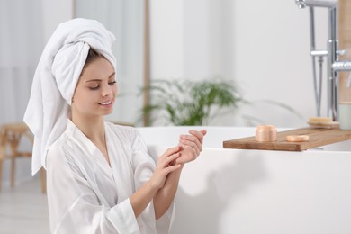 Beautiful happy woman in white robe applying cream near tub in bathroom