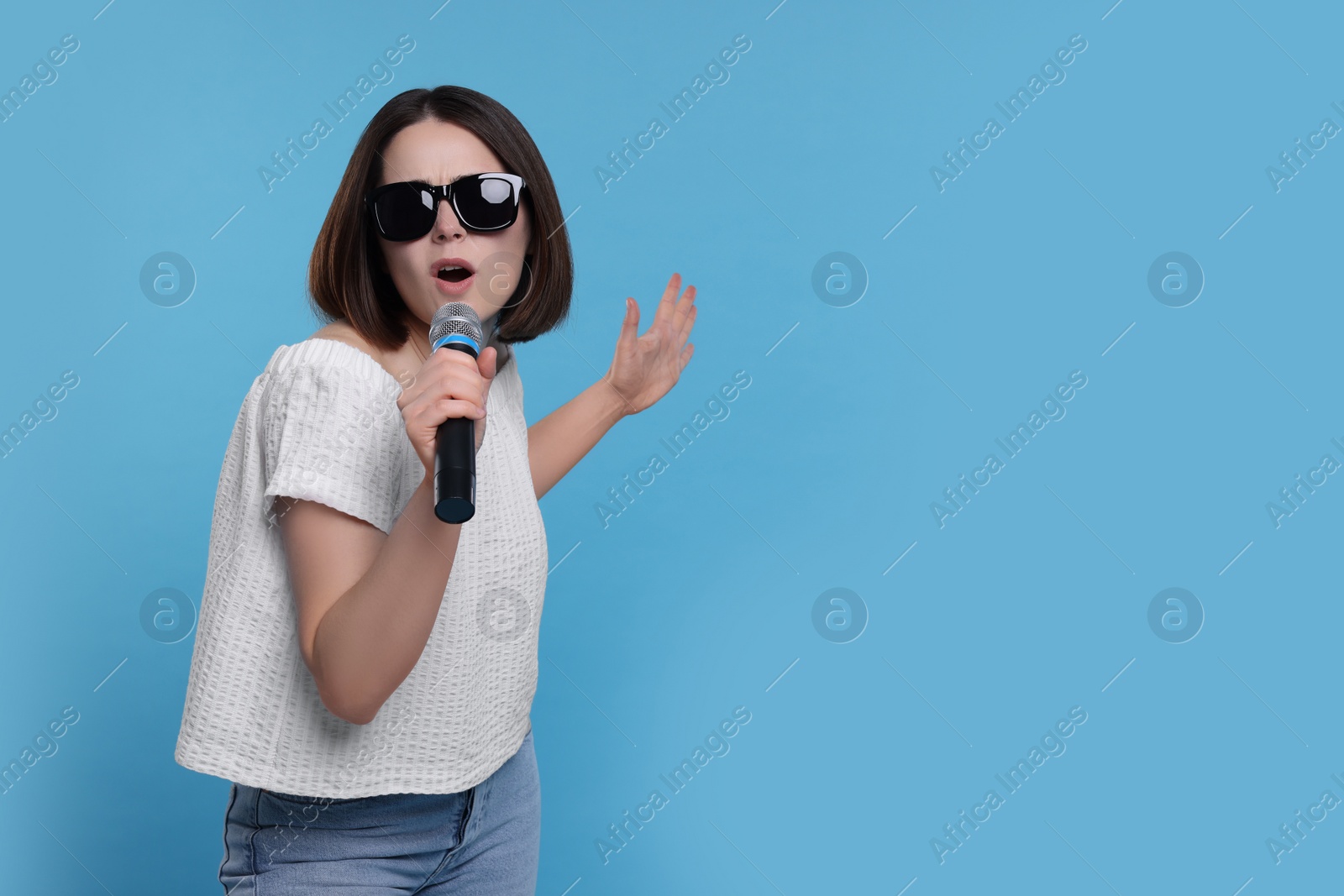 Photo of Beautiful young woman with sunglasses and microphone singing on light blue background, space for text