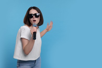 Beautiful young woman with sunglasses and microphone singing on light blue background, space for text