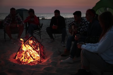 Photo of Group of friends gathering around bonfire on beach in evening. Camping season