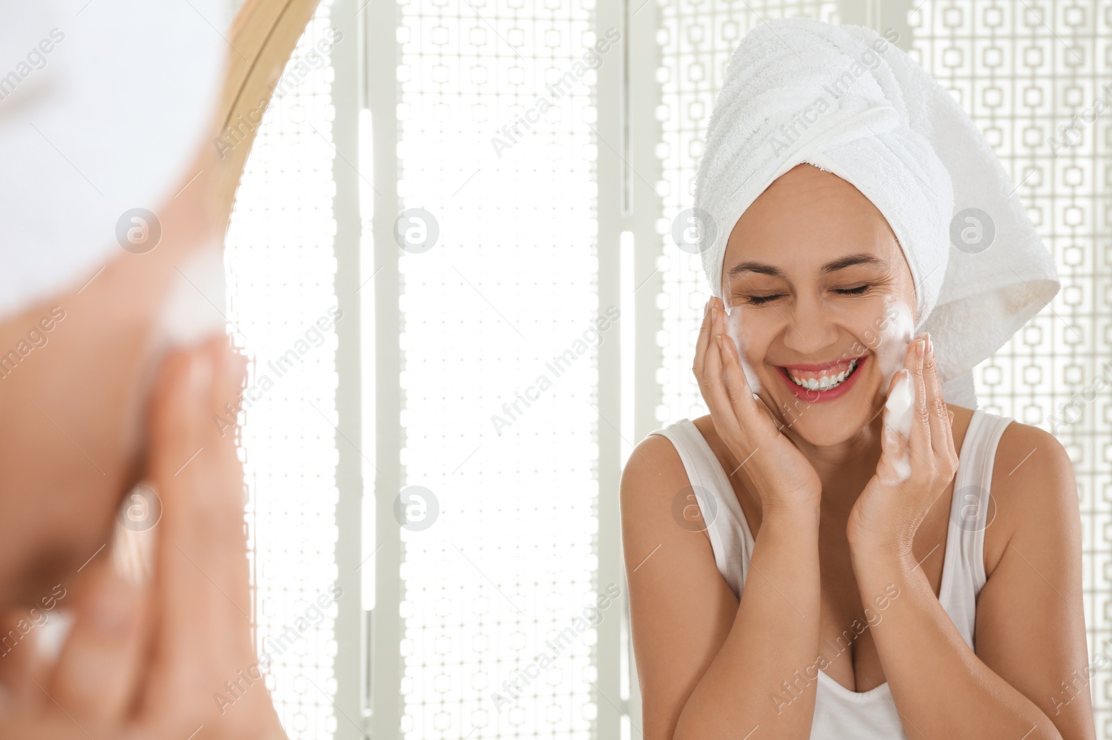 Photo of Happy mature woman applying cleansing foam onto face near mirror in bathroom