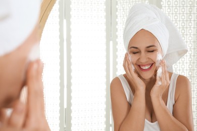 Photo of Happy mature woman applying cleansing foam onto face near mirror in bathroom