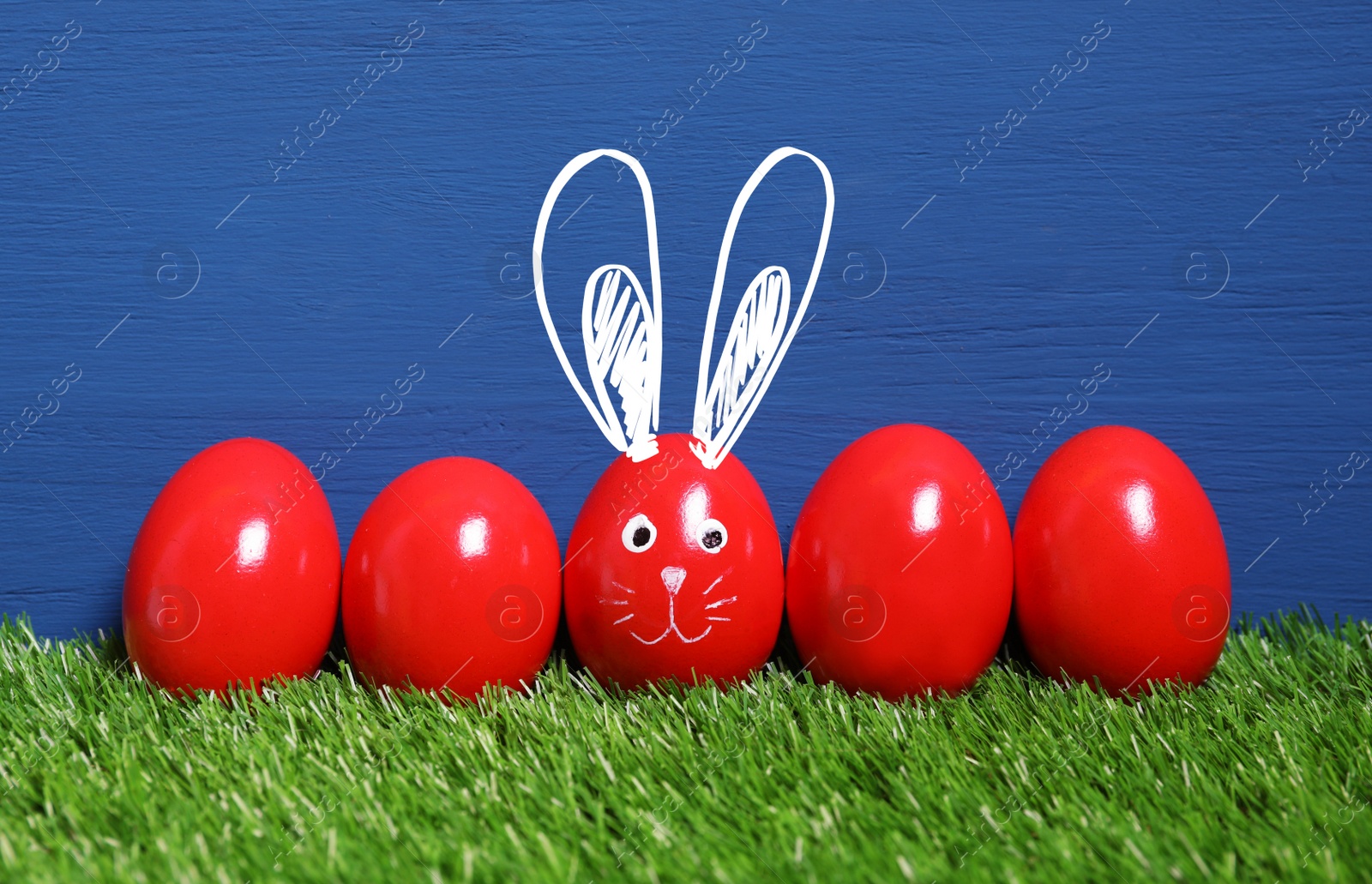 Image of One egg with drawn face and ears as Easter bunny among others on green grass against blue wooden background